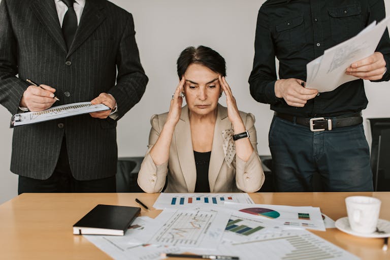 A stressed businesswoman holding her head with colleagues holding documents in a corporate office setting.