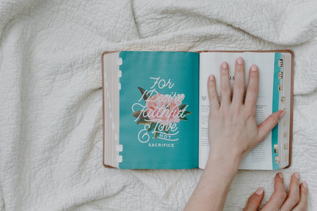 An open Bible on a white quilt with a hand resting on the page, promoting faith and reflection.